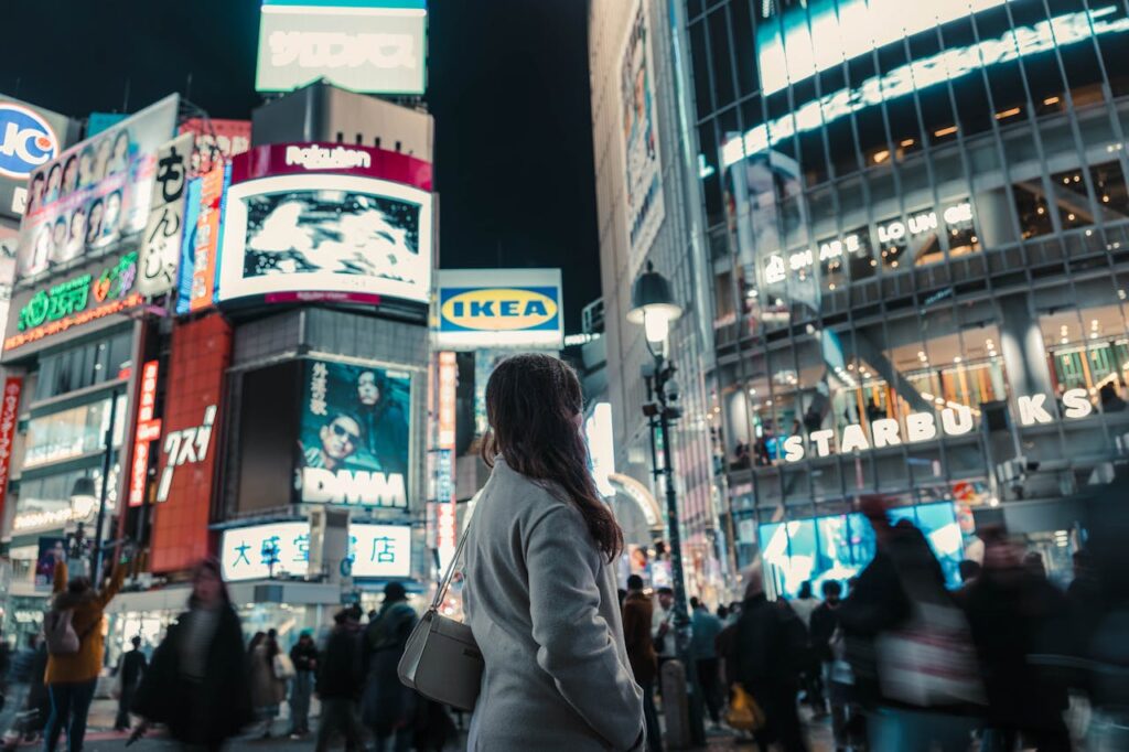 A bustling evening in Shibuya, Tokyo with bright neon signs, lively crowds, and a lone woman gazing at the cityscape.